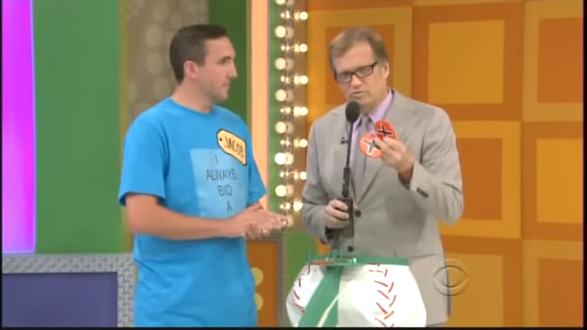 A Price is Right contestant wearing a blue shirt with the name 'Jacob' stands next to Drew Carey, who is holding a strike chip, while a baseball-themed Three Strikes game setup is visible in front of them.