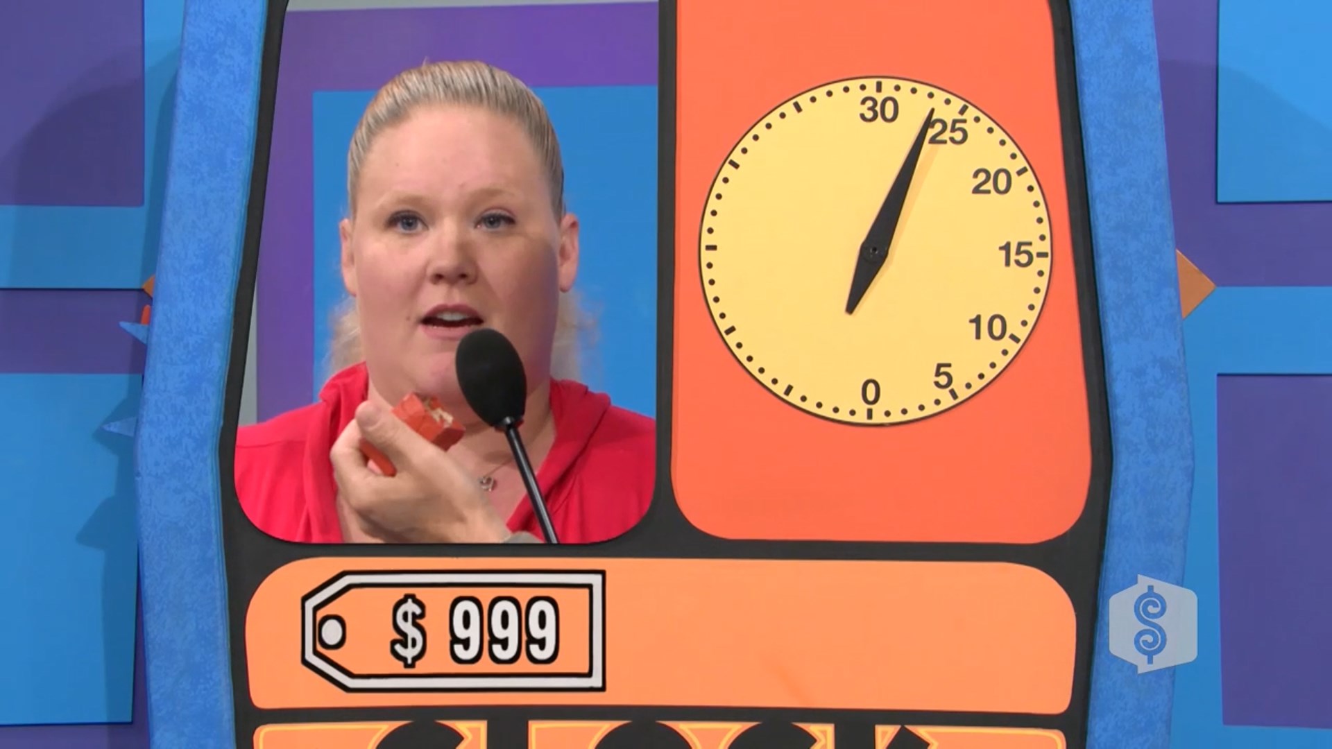 A contestant in the Clock Game holds a bidding paddle while speaking into a microphone, with a large countdown clock showing approximately 25 seconds remaining and a price tag reading 9.
