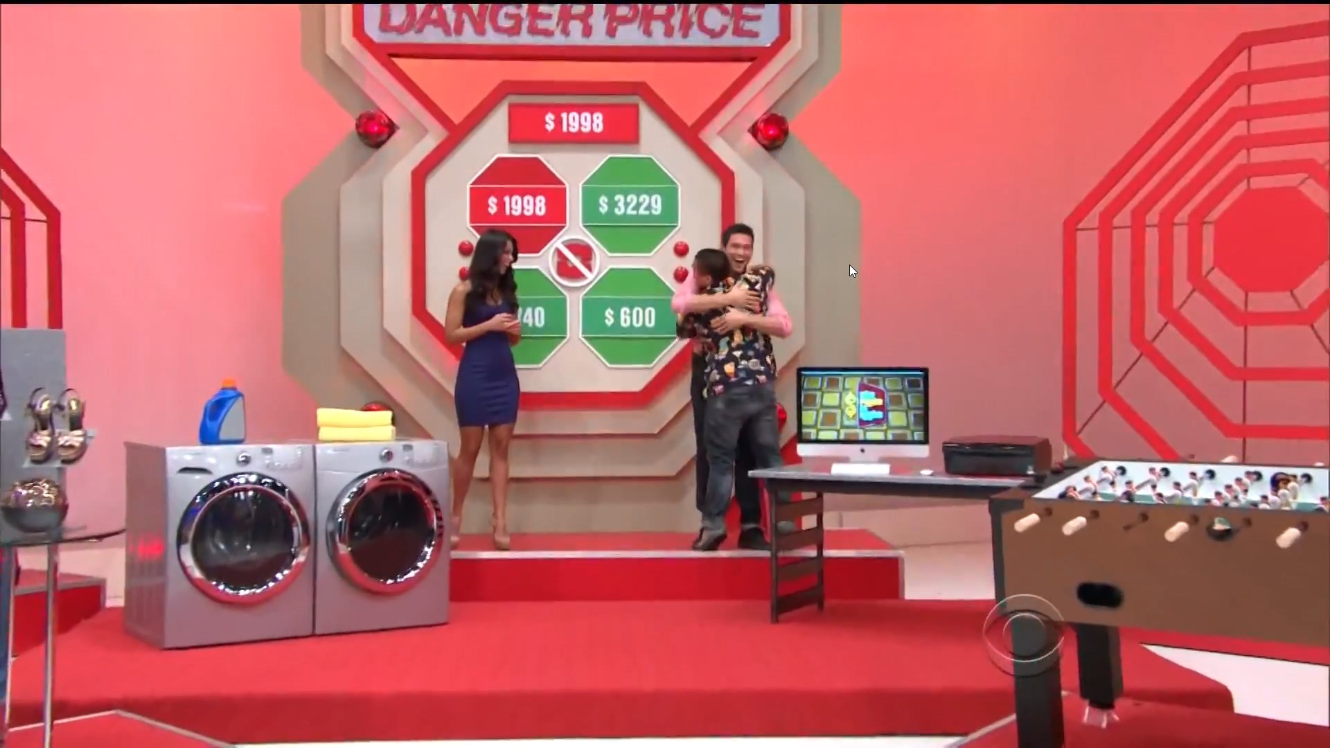 Contestants celebrate while standing next to a game board for 'Danger Price,' which displays various prize values including 98, 29, 0, and 0, alongside two washing machines and a foosball table.