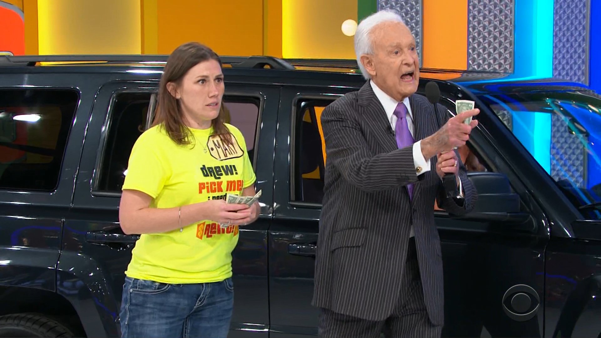 Bob Barker enthusiastically interacts with a contestant holding cash, while a black car is visible in the background.