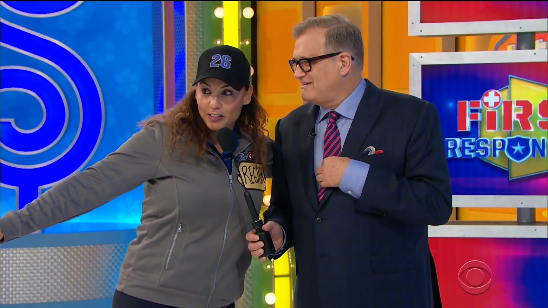 Contestant Regina gestures while speaking to host Drew Carey on the set of The Price Is Right, with colorful game show backdrop.