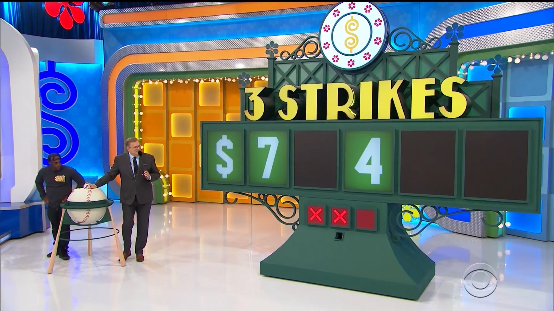 A game board for 'Three Strikes' featuring the numbers , _, 4 displayed in a colorful set on 'The Price is Right', with a contestant and host interacting beside a large baseball prop.