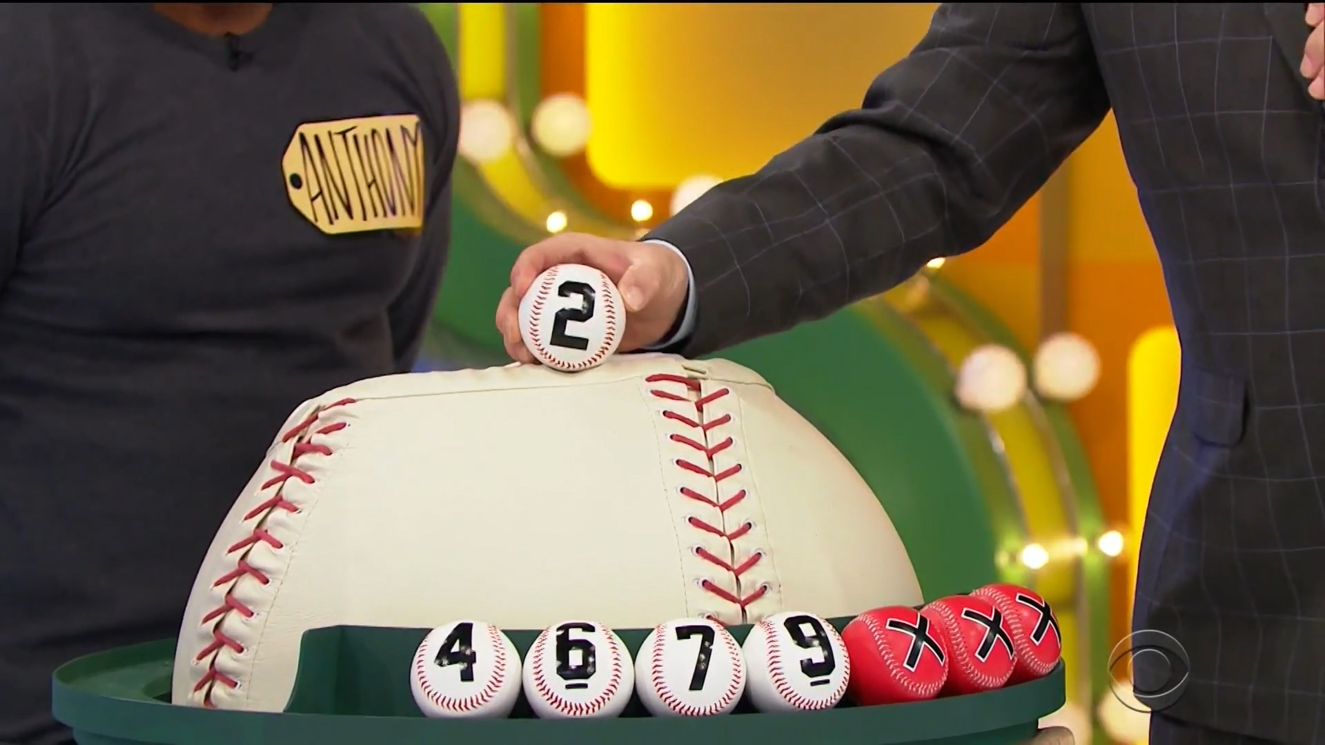 A host holds a baseball with the number 2 while preparing to place it on a large baseball prop that represents the game Three Strikes. Below, several baseballs with the numbers 4, 6, 7, and 9 are displayed, alongside red strike balls.