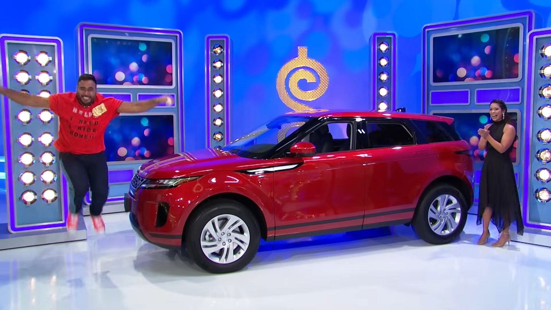 A contestant joyfully jumps next to a red SUV showcased on stage at a game show, while a female host applauds beside him.