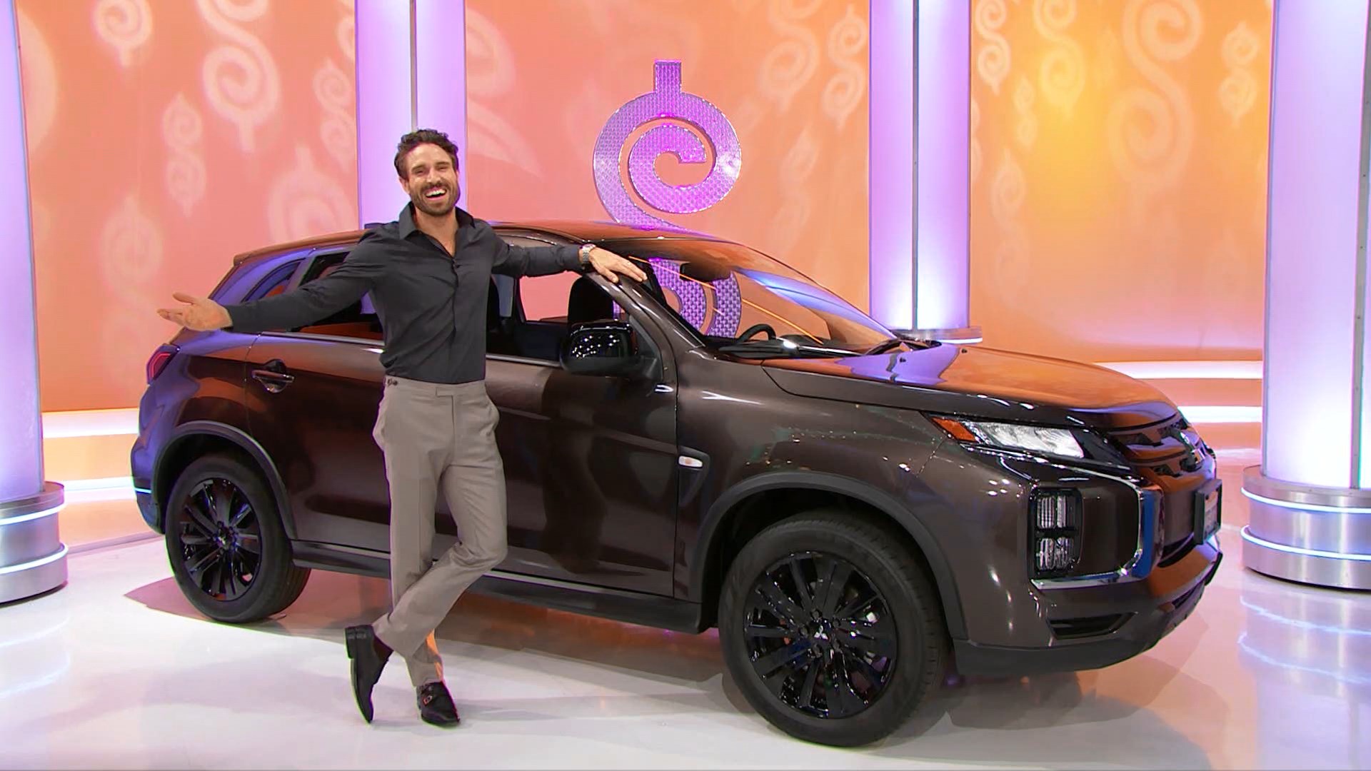 A model enthusiastically poses by a brown SUV on a game show set, with decorative columns in the background.