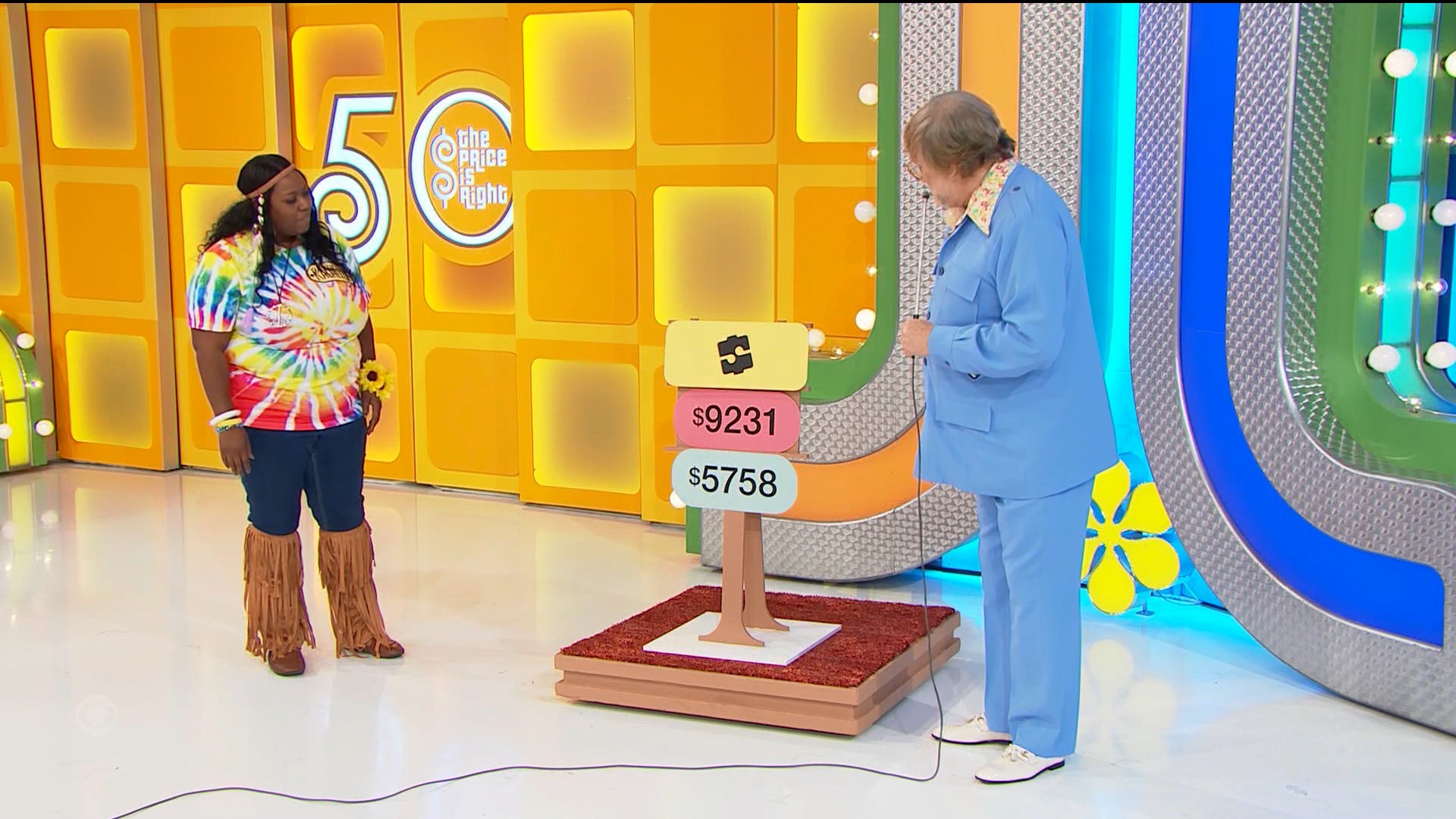 A contestant wearing a colorful tie-dye shirt and fringe pants stands next to a pricing game display on 'The Price is Right,' which shows two price tags: 31 and 58.