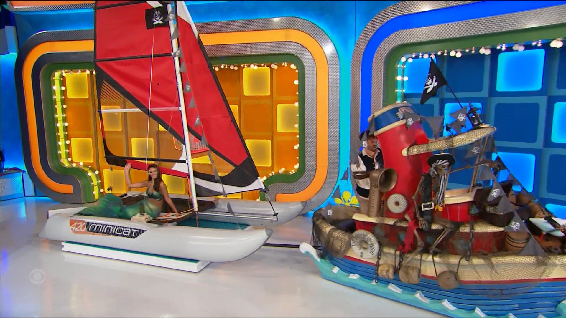 A game show setting featuring two boats: a sleek white sailboat with a red sail on the left, and a pirate-themed boat on the right, complete with flags and decorations. A contestant poses on the sailboat while a host engages with the audience.