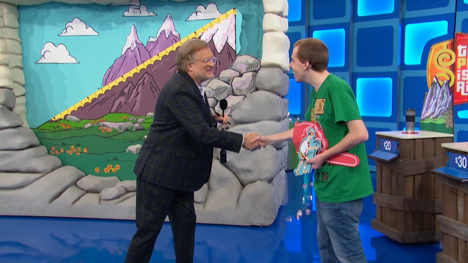 A contestant shakes hands with the host of The Price is Right in front of a colorful mountain backdrop representing the Cliff Hangers game.