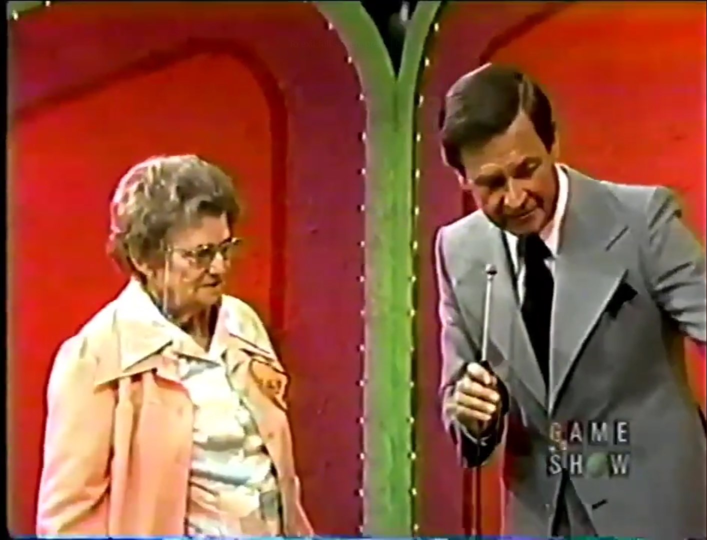 A contestant and Bob Barker stand together, with the host holding a microphone and gesturing while explaining the game rules.
