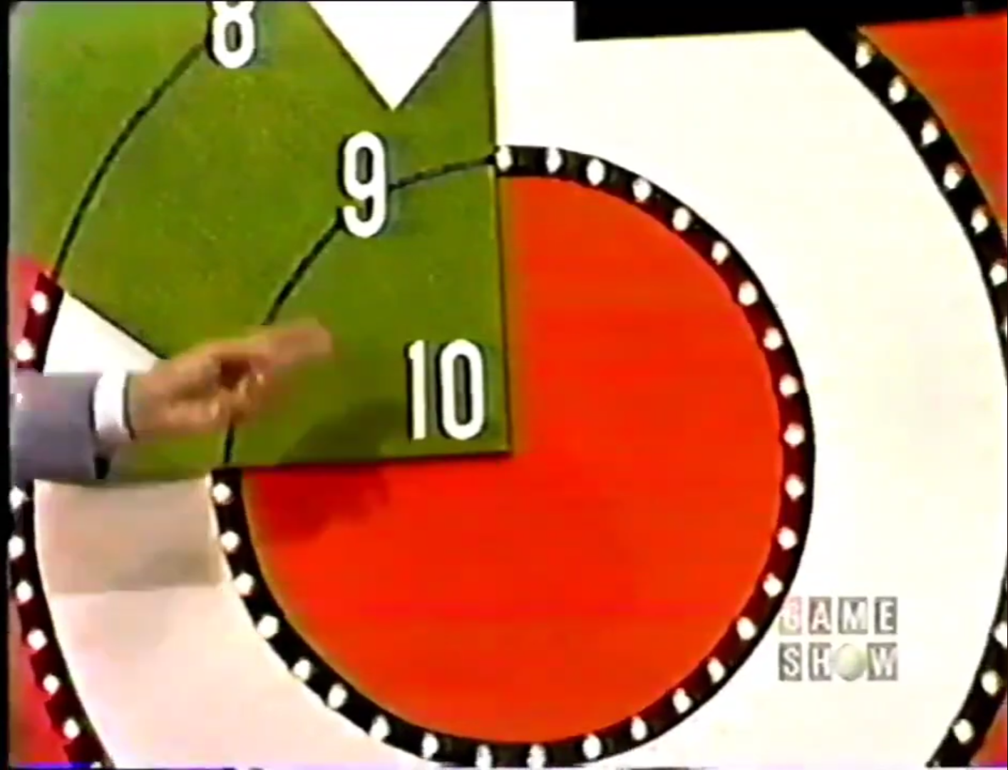 A hand pointing at the bullseye target displaying numbers 8, 9, and 10 in a game show setting.