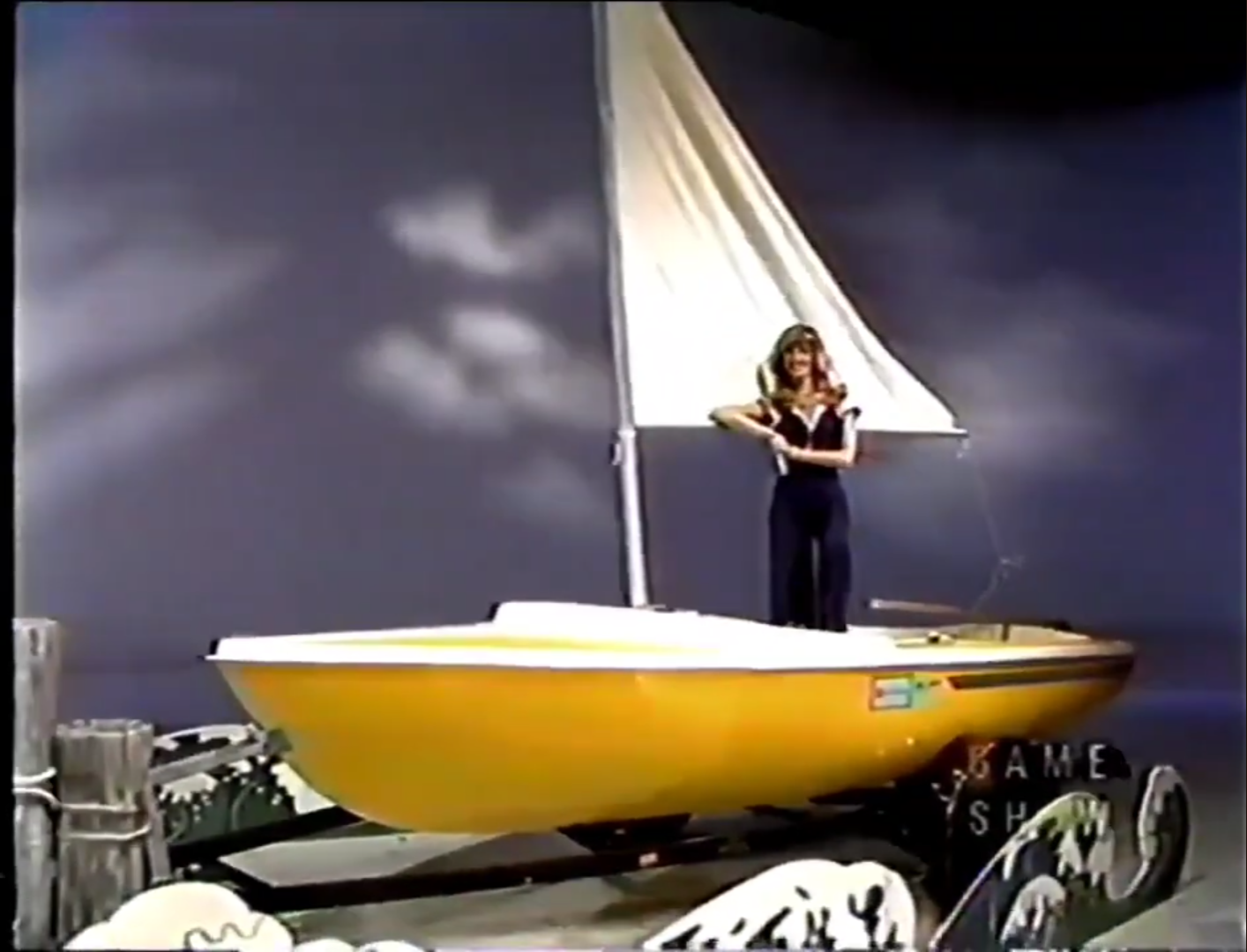 A woman stands on a yellow sailboat on a game show set, with a blue-grey background and wave decorations at the base.