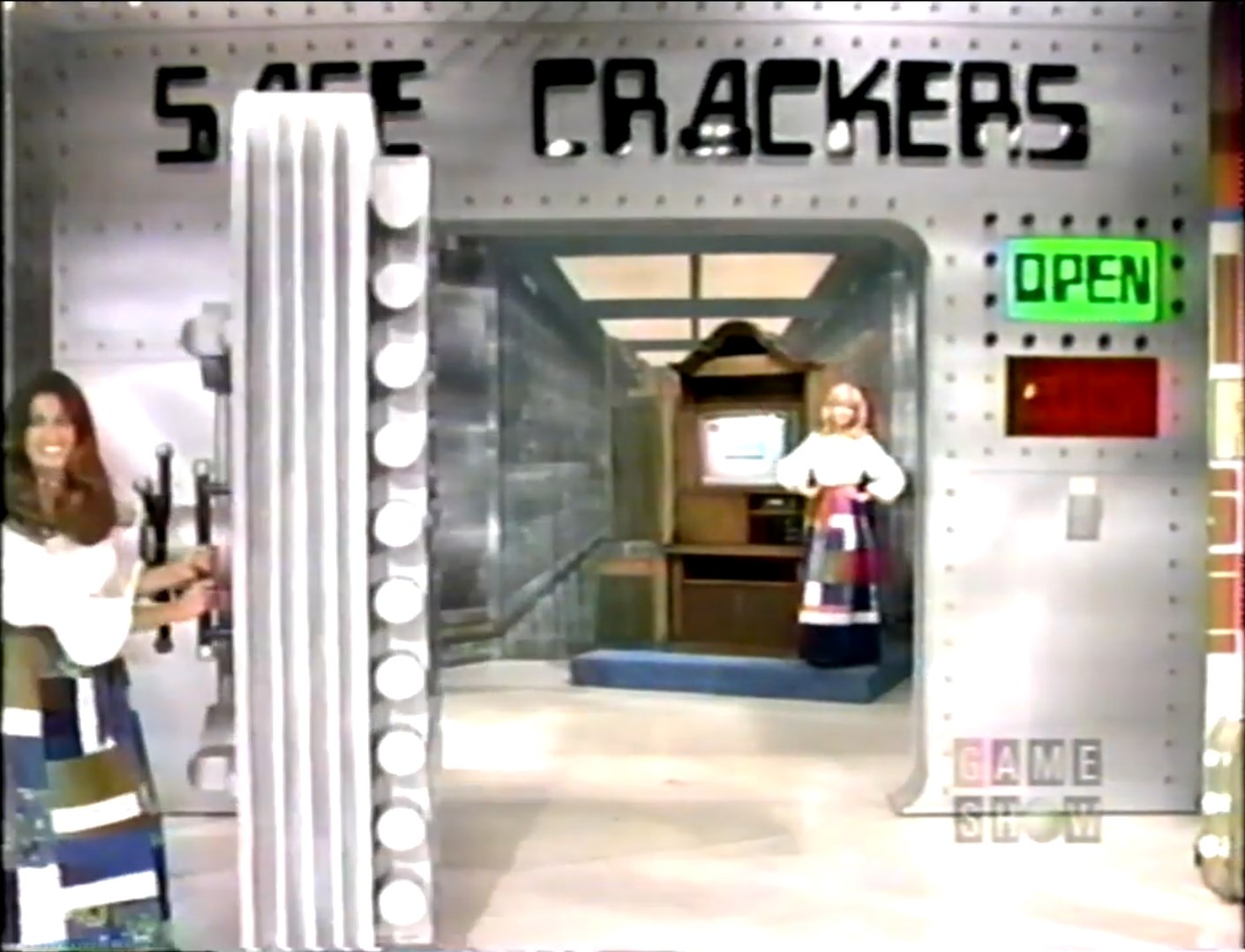 A scene from the 'Safe Crackers' game on The Price is Right featuring a large safe with an 'OPEN' sign. A female model stands near the safe opening, while another woman is seen behind the safe, dressed in a colorful outfit.