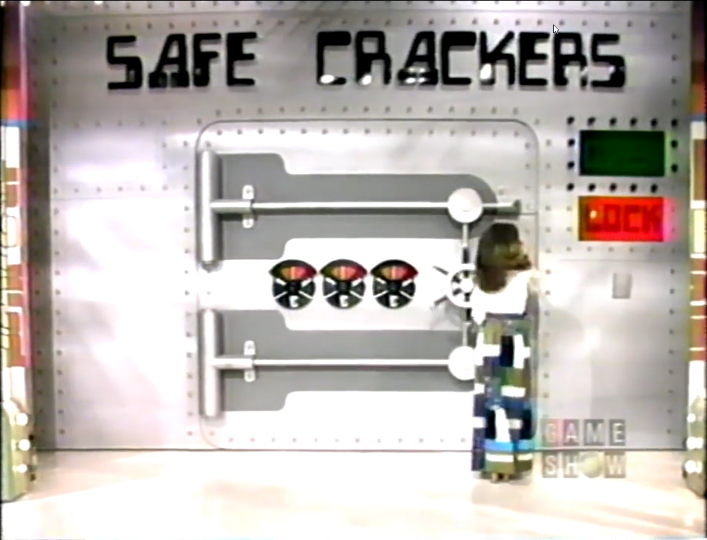 A model locking a large safe marked 'SAFE CRACKERS' on a game show set, attempting to guess the combination.