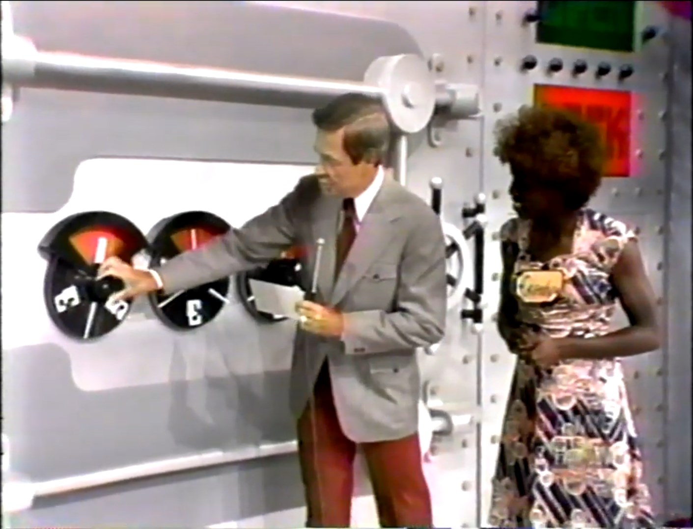 A contestant and the host interact with a combination lock as part of the Safe Crackers game, featuring three dials displaying numbers.