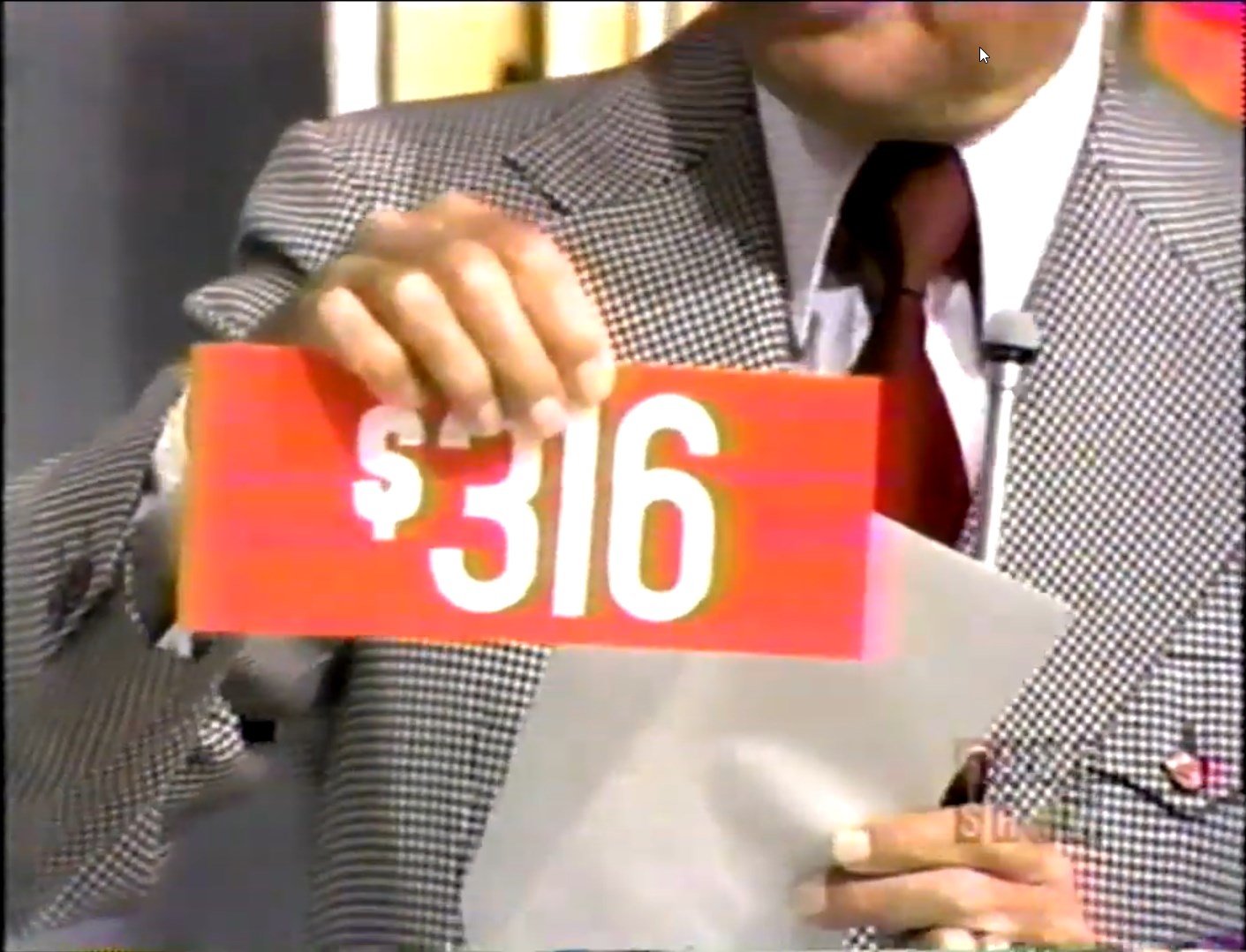 6 price displayed on a red card held by a host, with a checkered suit in the background.