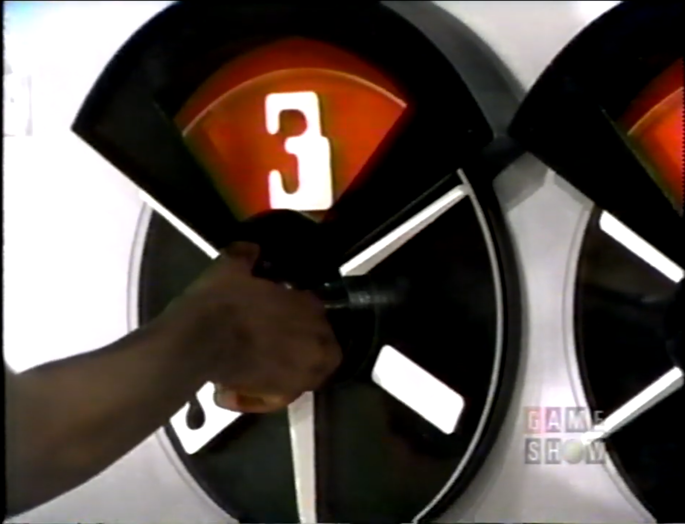 A close-up image of a contestant's hand turning the dial on a game show wheel, showing the number 3.