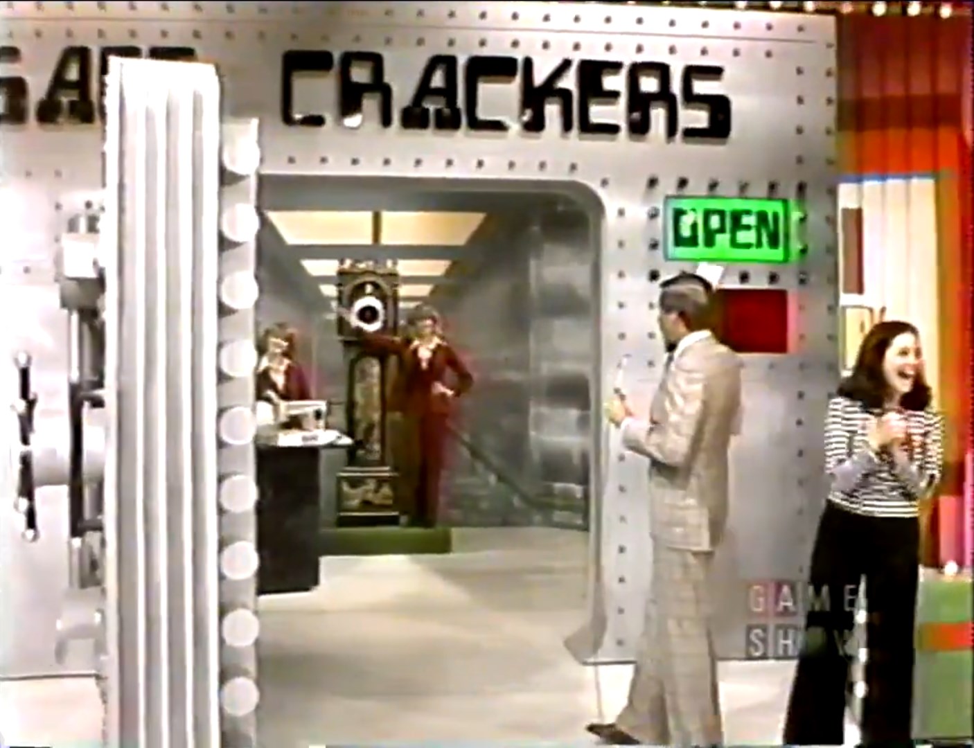 Contestants and host in front of the game show set for 'Safe Crackers,' featuring a large safe with an 'OPEN' sign.