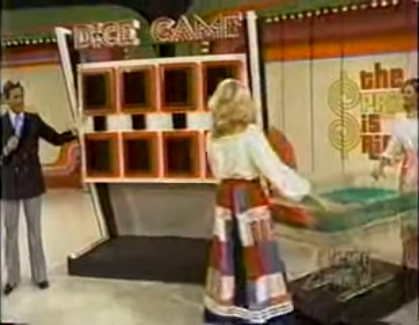 A contestant plays the Dice Game on stage, rolling oversized dice in front of a game board with empty slots and a colorful background.