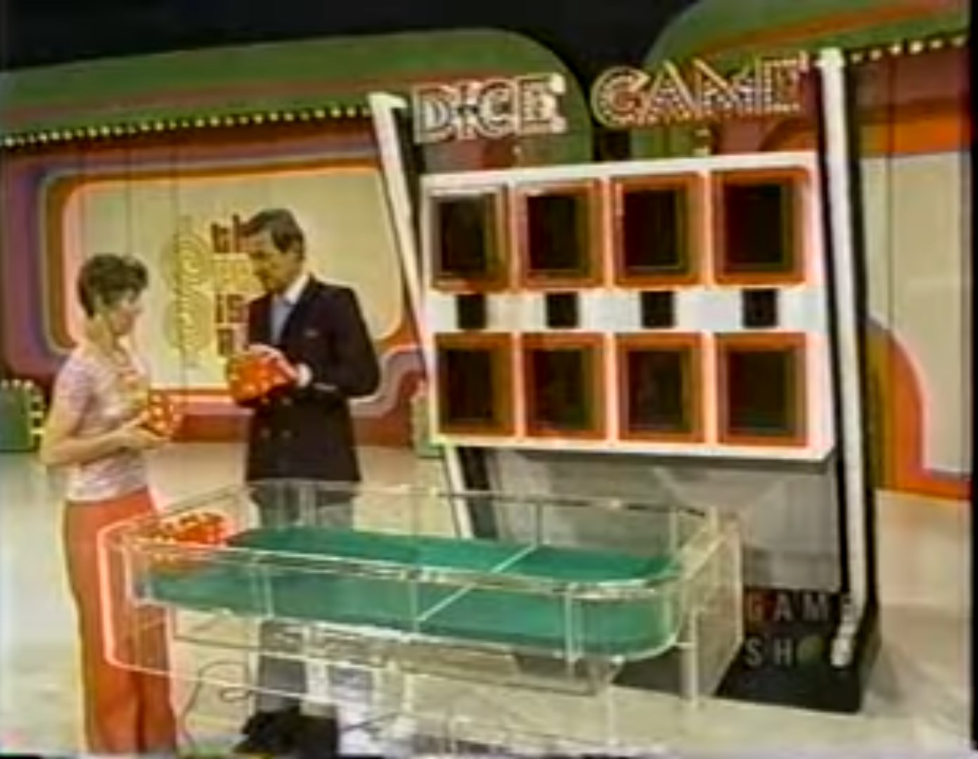A contestant holds oversized dice while a host stands beside her, with a game board labeled 'DICE GAME' in the background.
