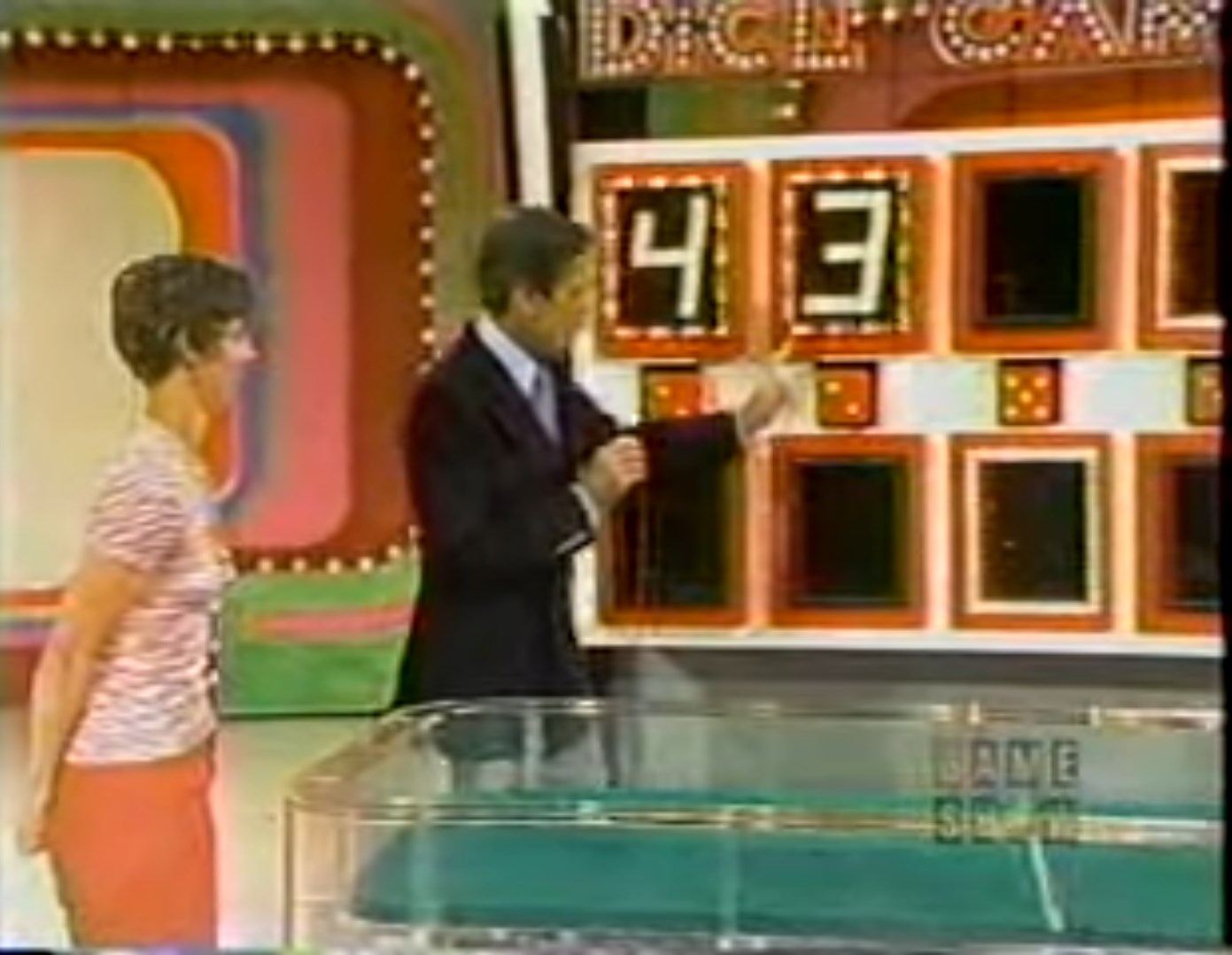 A contestant stands beside a game show host while they interact in front of a brightly colored game board displaying the numbers 4 and 3.