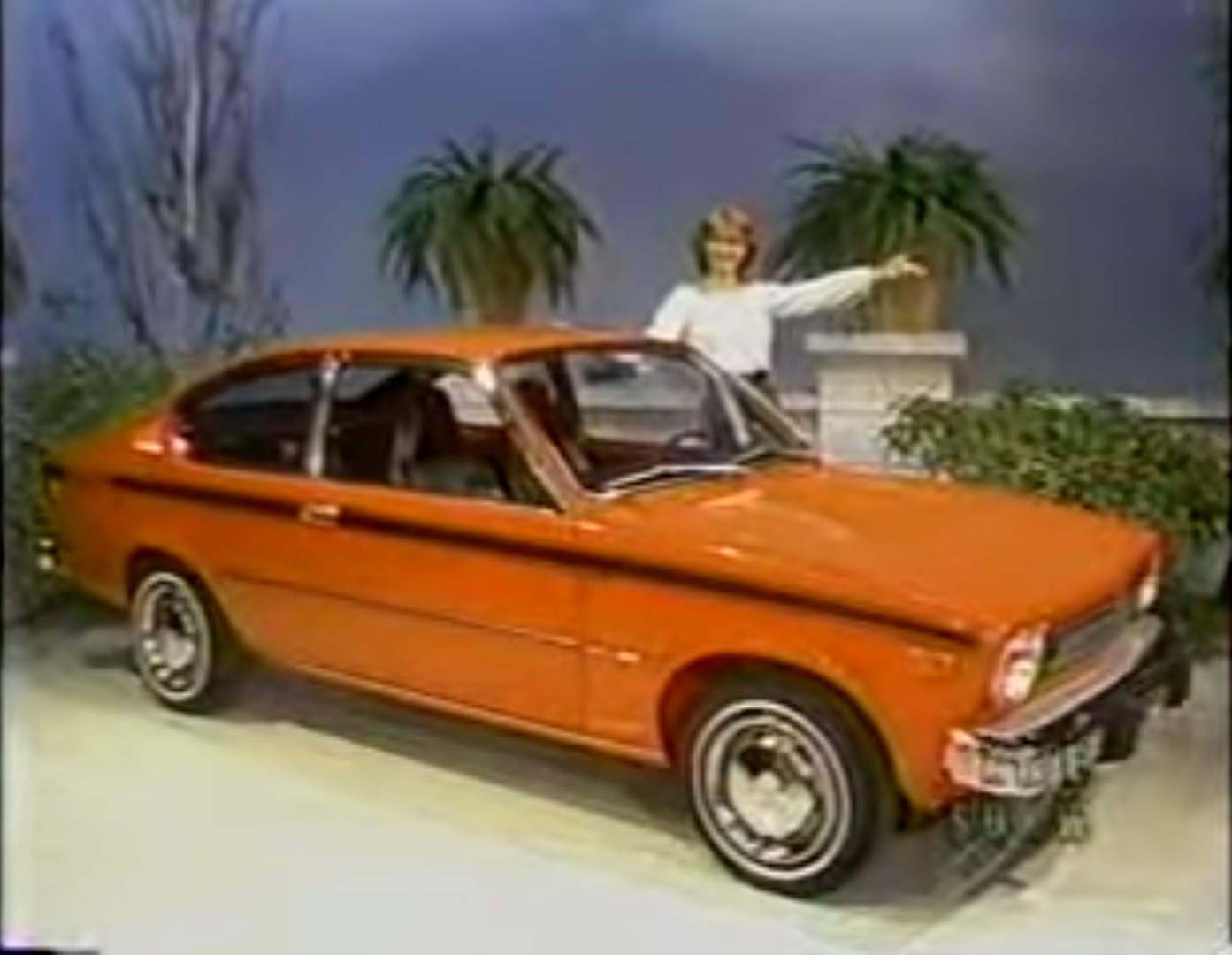 A model stands next to an orange car, pointing at it with excitement during the Dice Game segment.