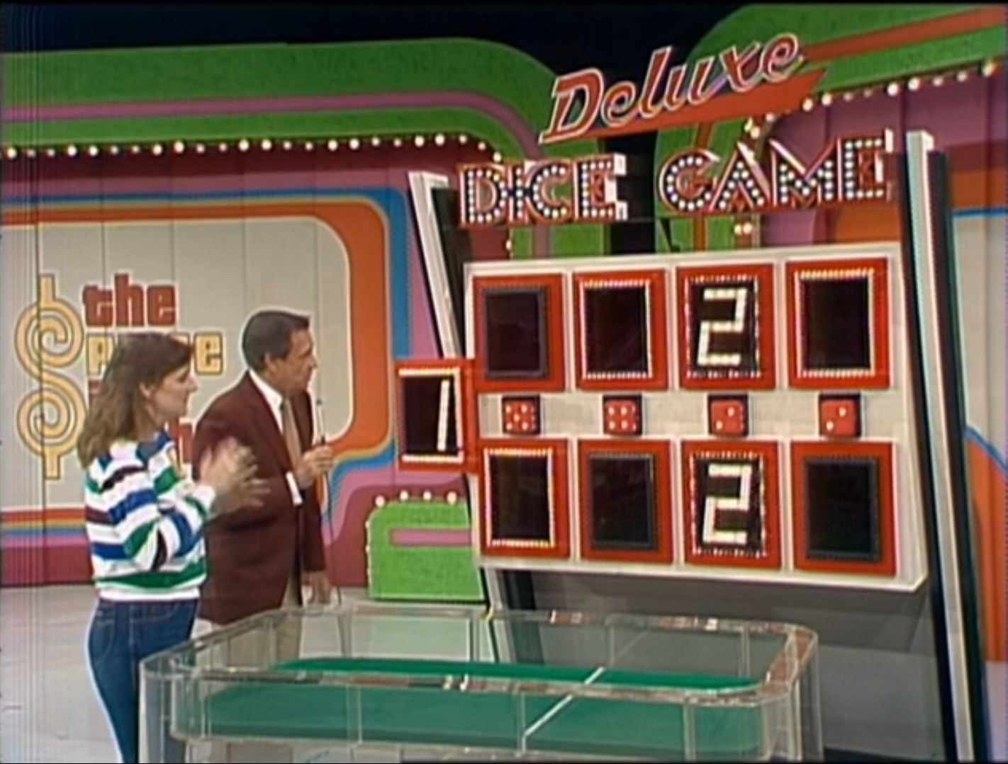 A contestant and host stand beside the Deluxe Dice Game board, featuring numbered slots and giant dice, during a game show.