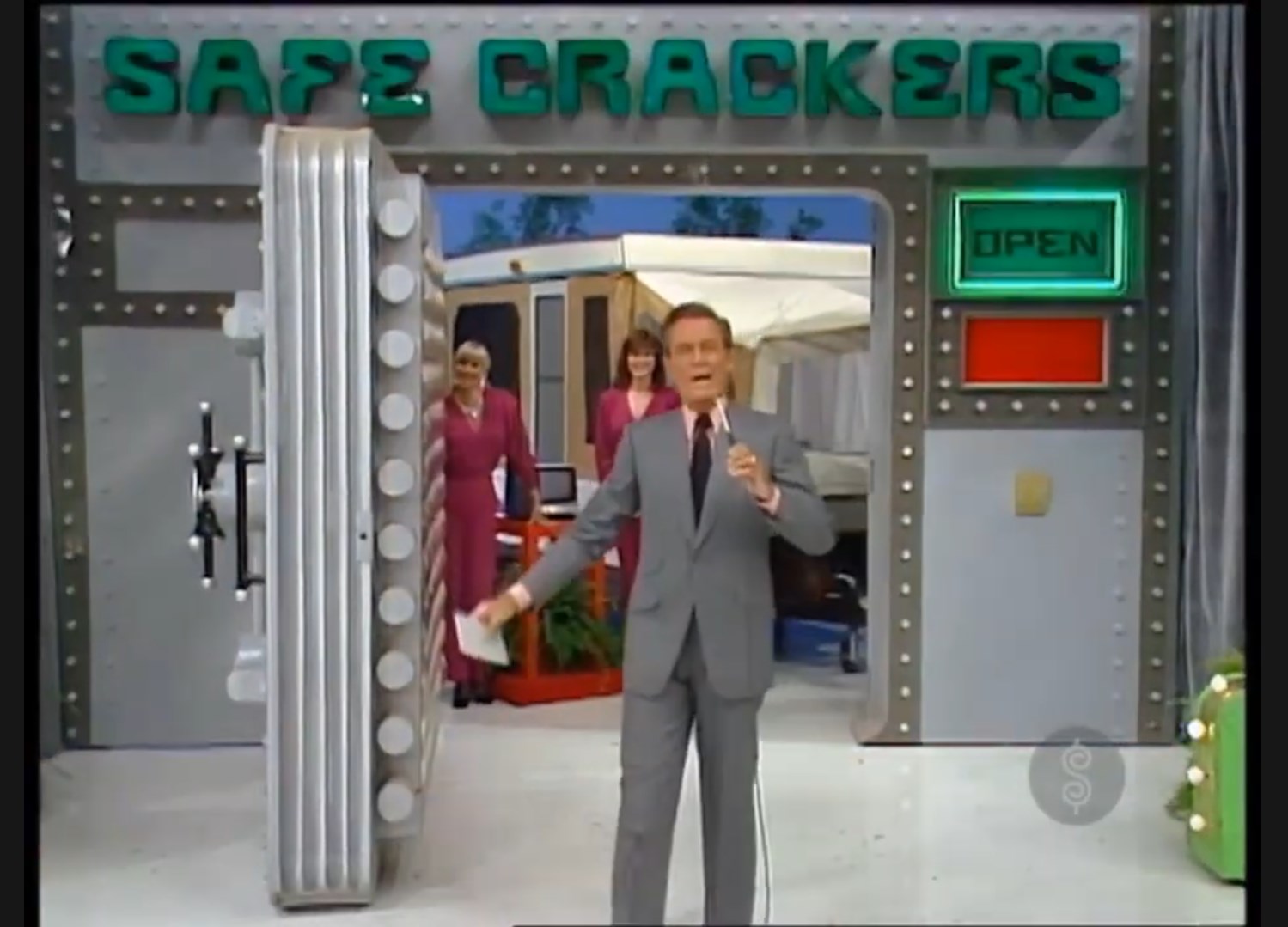 A game show host stands in front of a large safe labeled 'SAFE CRACKERS,' with an open door revealing prizes inside. Two female contestants watch in the background.