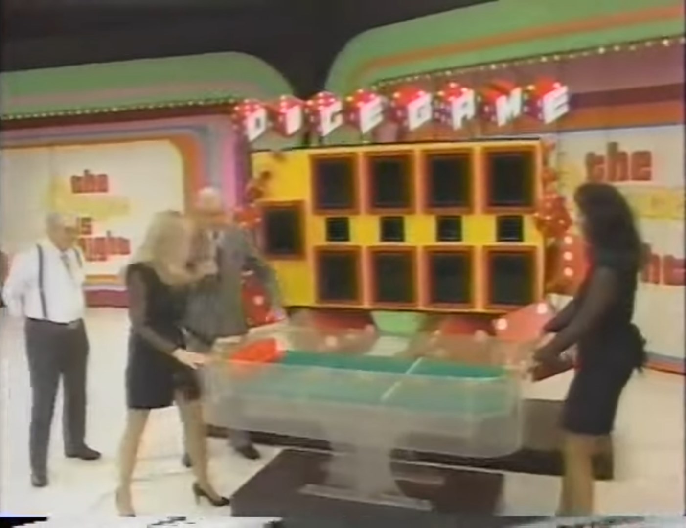 A group of contestants assists in setting up the Dice Game board, featuring an oversized transparent table and a colorful backdrop.