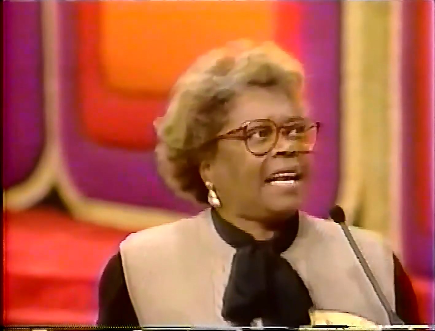 A contestant passionately speaking into a microphone during the Dice Game segment, wearing glasses and a stylish outfit, against a colorful game show backdrop.