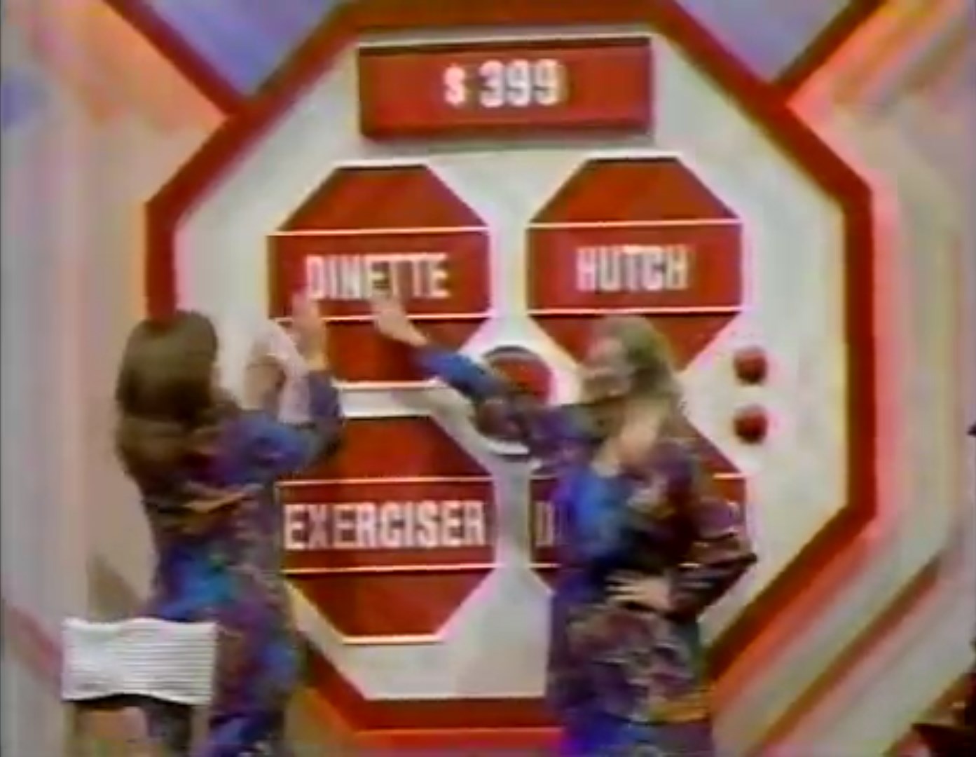 Two contestants are reaching for the prizes displayed on a game board for the Danger Price game, which features a color display with the words 'DINETTE', 'HUTCH', and 'EXERCISER' alongside a Danger Price of 9.