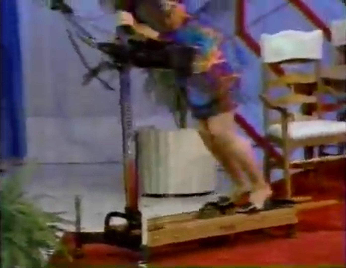 A contestant demonstrating an exercise machine on stage, struggling to maintain balance while using it.
