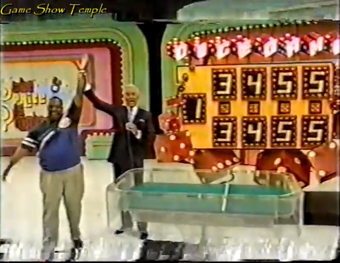 A contestant celebrates while holding their arm up in victory next to a host, with a game board displaying the numbers 3455 in the background.