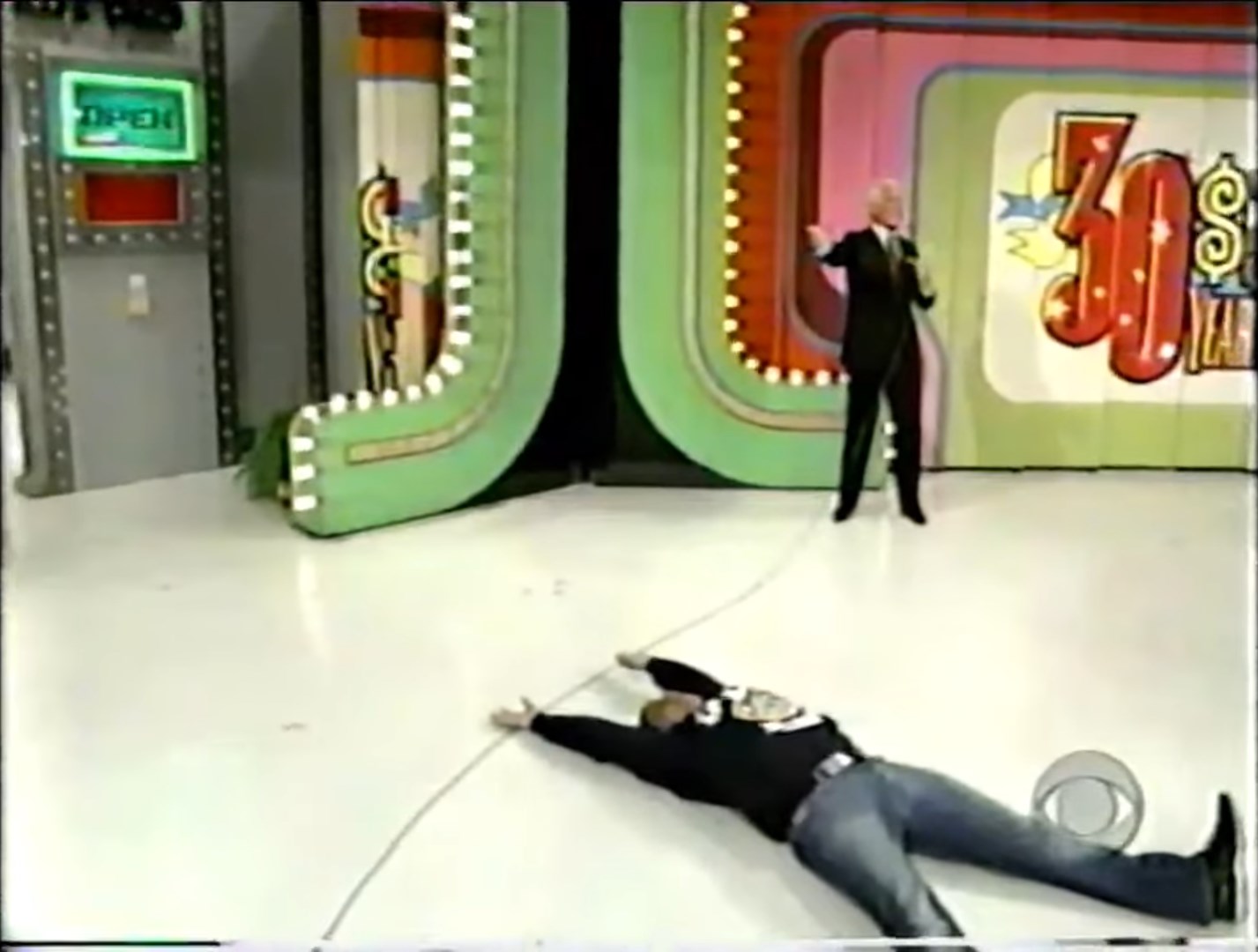 A contestant lies on the floor while a host gestures during a game on a colorful stage set.