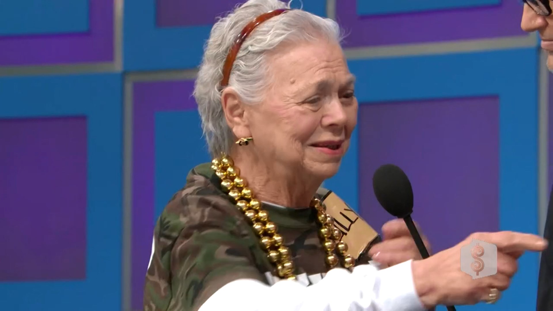 An elderly woman with short gray hair, wearing a camouflage shirt and gold bead necklace, speaks into a microphone while smiling on stage during a game show.