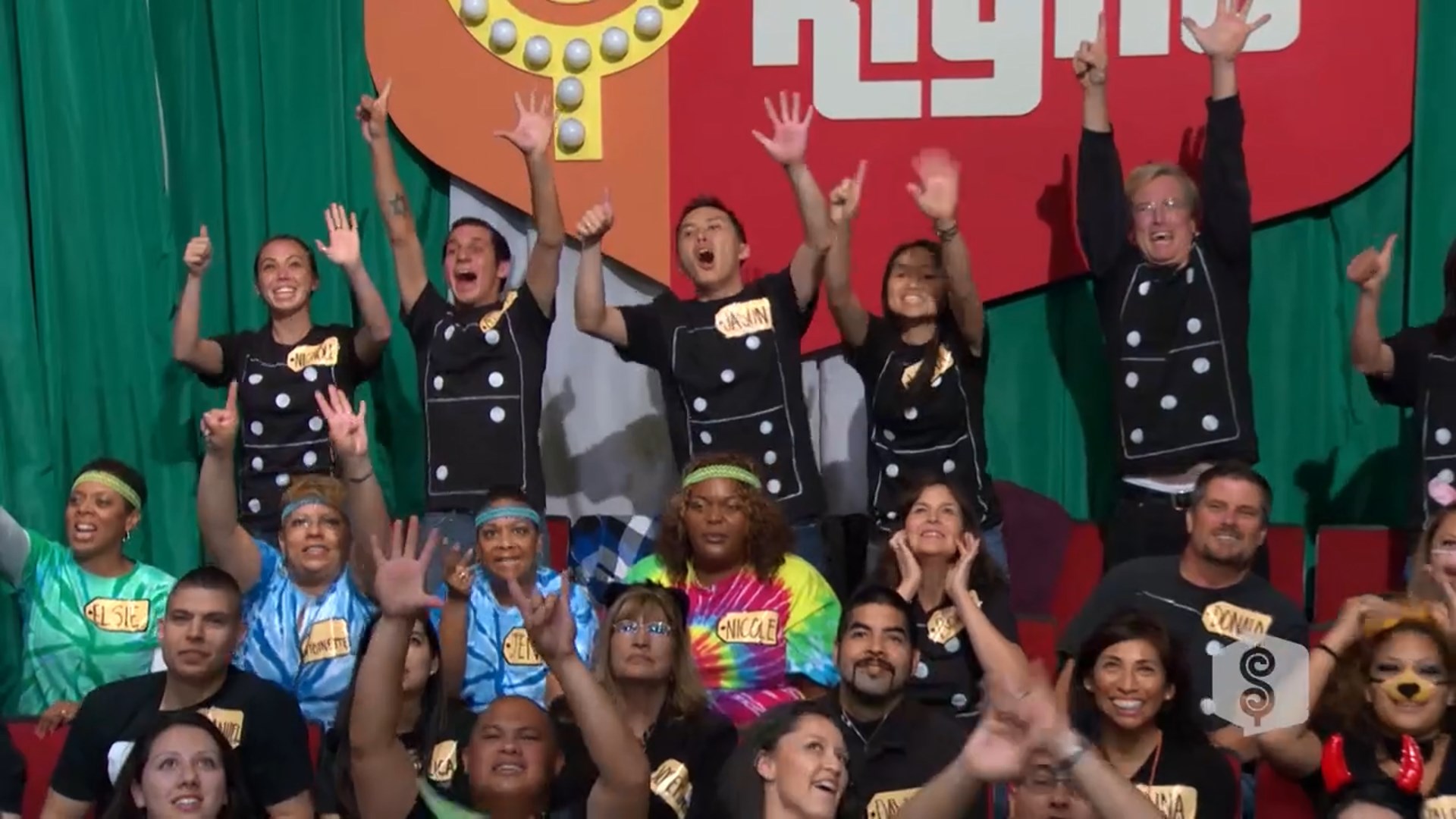 A lively audience cheering enthusiastically, wearing colorful shirts and matching name tags, with some making hand gestures and signs of excitement.