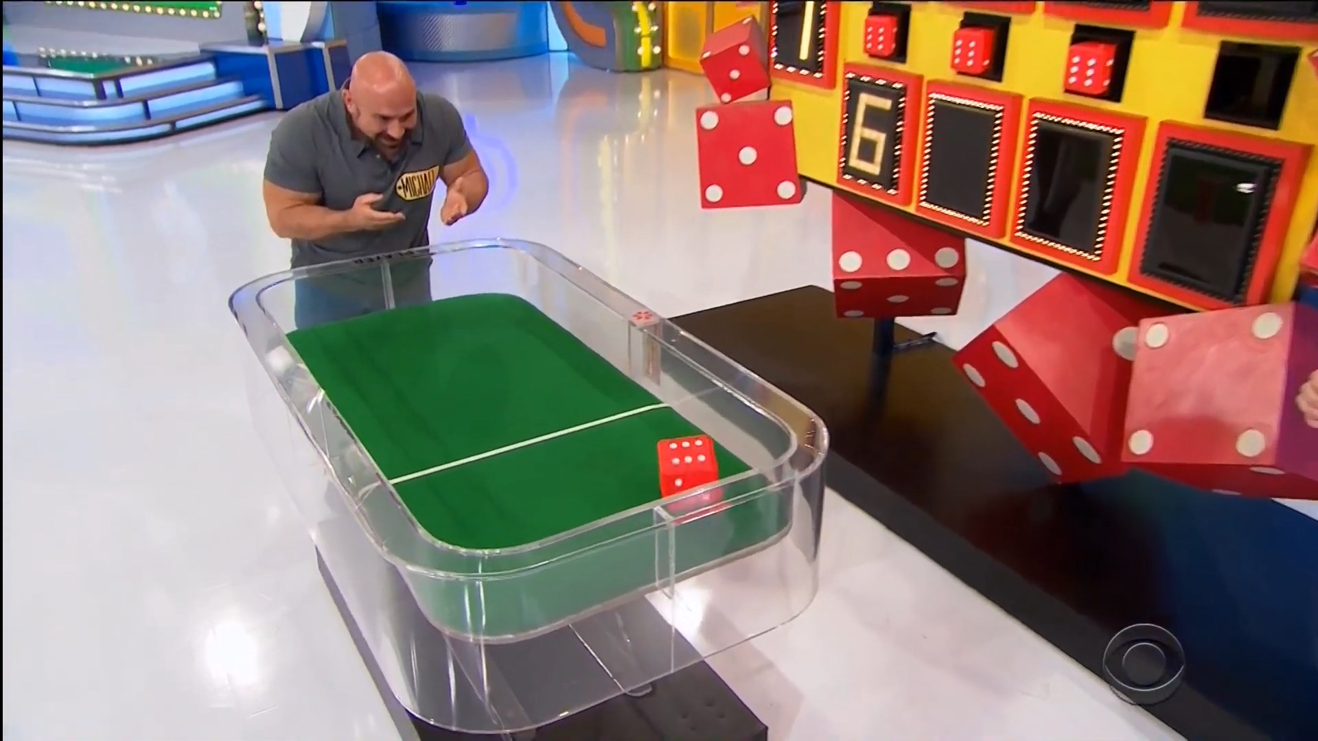 A contestant engages in the Dice Game, preparing to roll oversized dice on a green felt surface in a brightly lit studio.