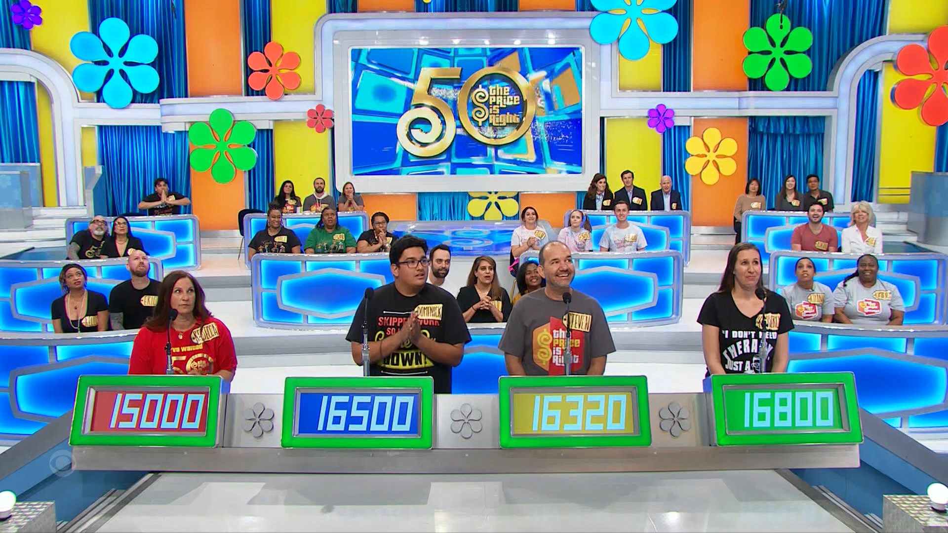 A group of contestants stands at a pricing game on The Price is Right, displaying their bids on illuminated boards, with a colorful backdrop featuring large flower designs.