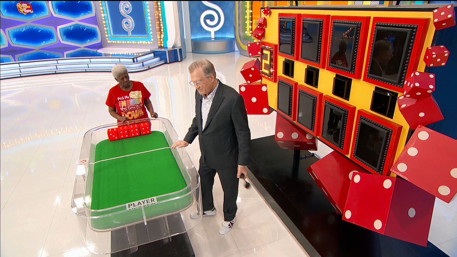 A contestant prepares to participate in the Dice Game, standing at a table with oversized dice, while Drew Carey explains the game mechanics and the colorful game board is visible in the background.