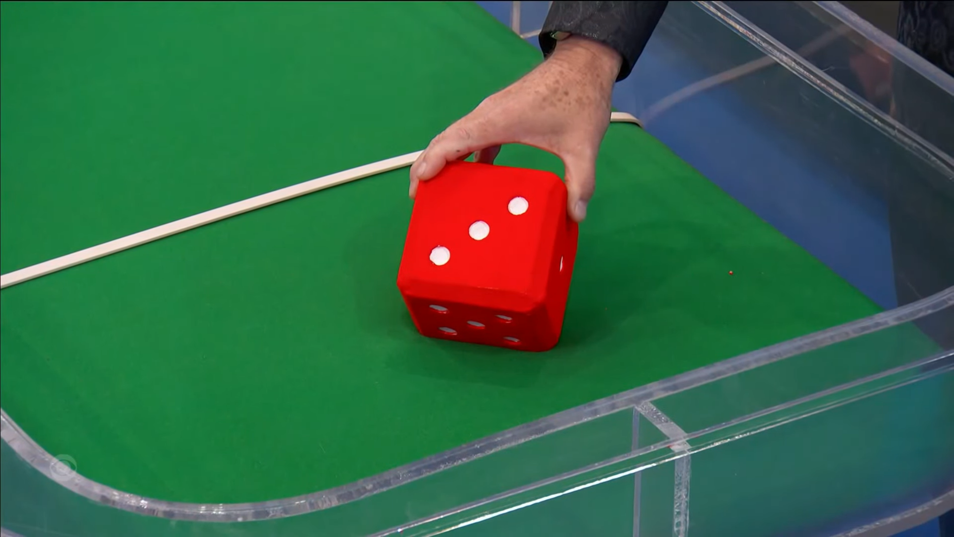 Drew Carey's hand holding a large red die with white dots, positioned above a green game board.