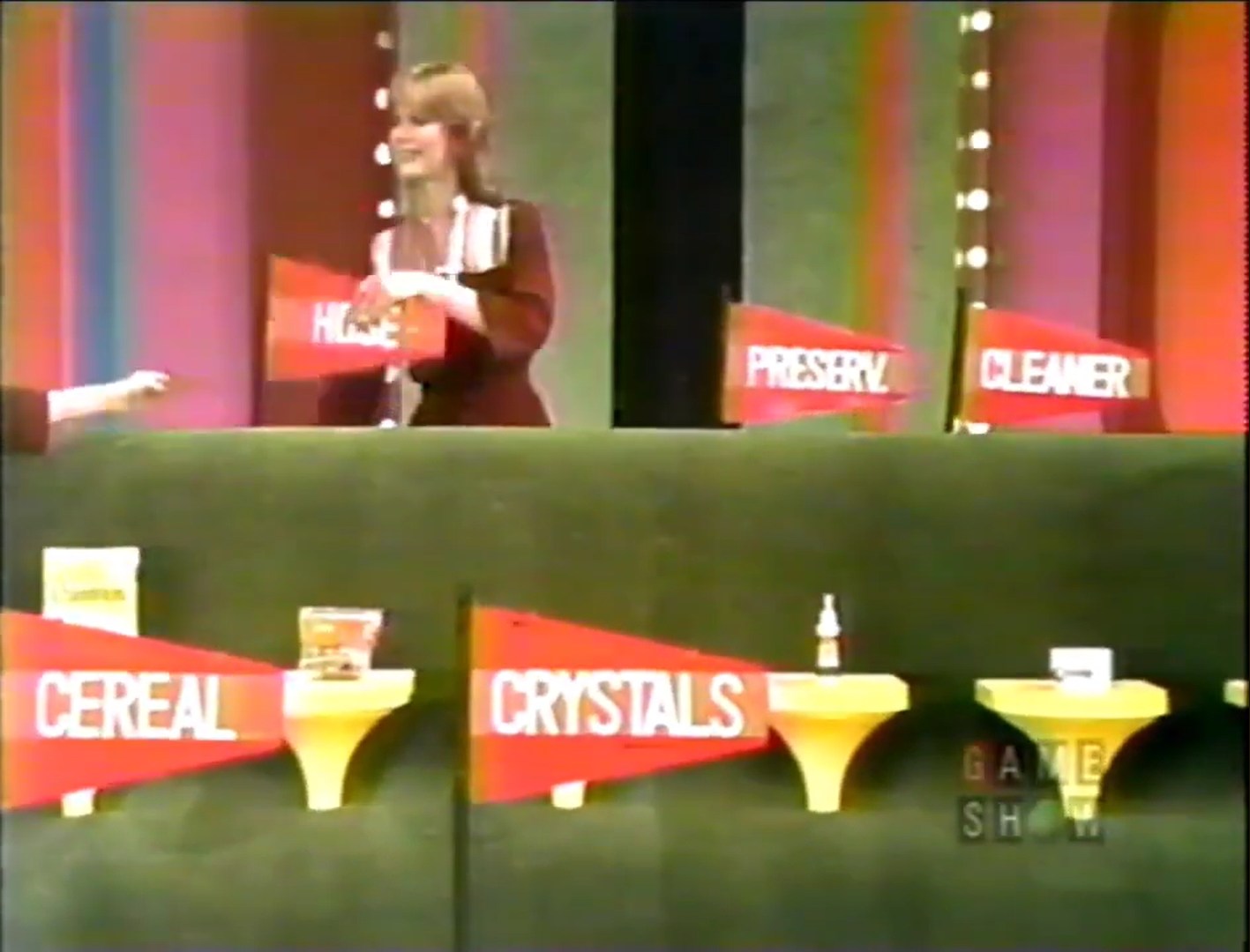 A model holding a flag labeled 'Hose' stands behind a green setup featuring grocery products with flags labeled 'Cereal', 'Crystals', 'Preserv.', and 'Cleaner' in a colorful game show environment.