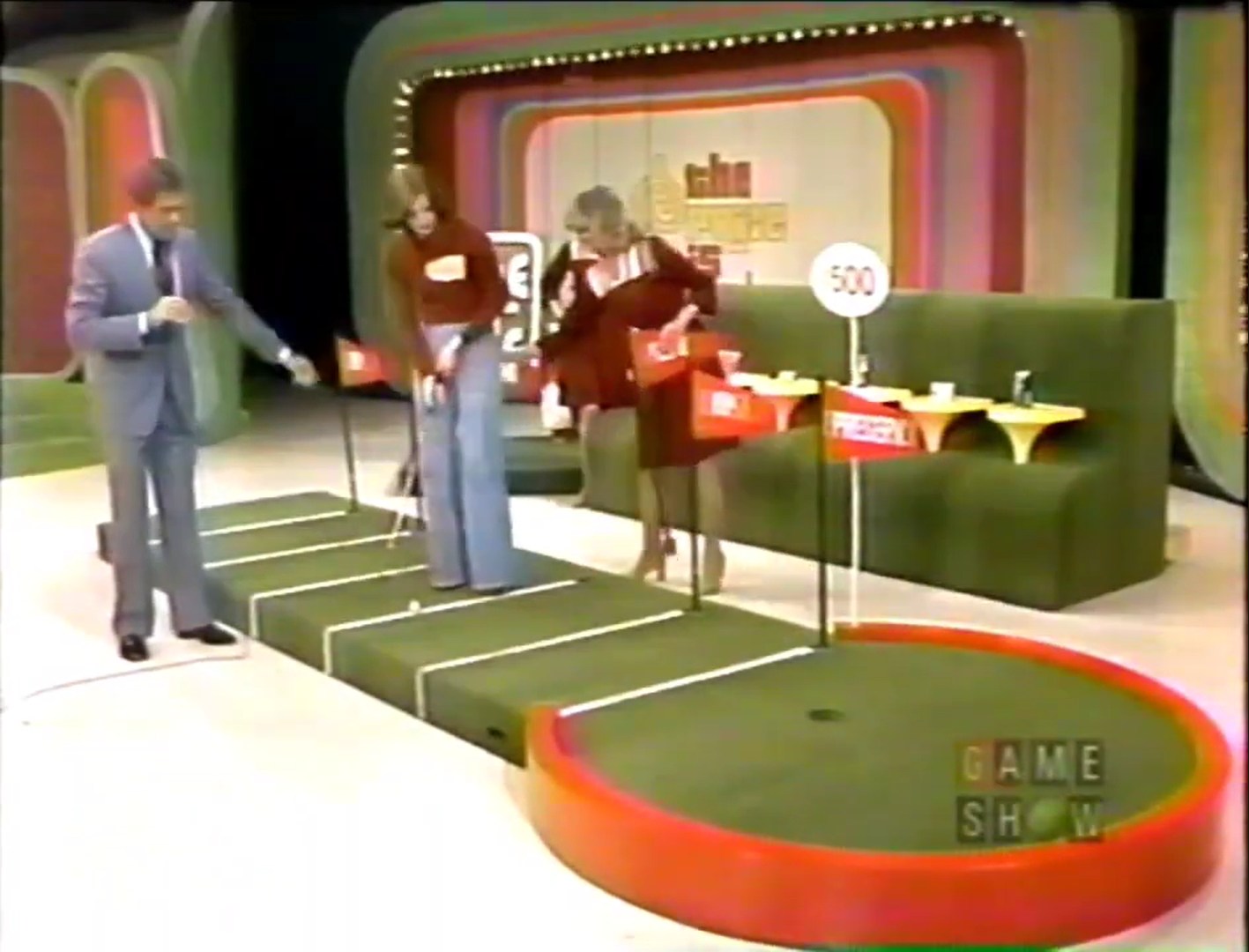 Contestants at a game show participate in the Hole in One game, with a putting green featuring multiple lines and flags. One contestant prepares to putt while the host gives instructions.