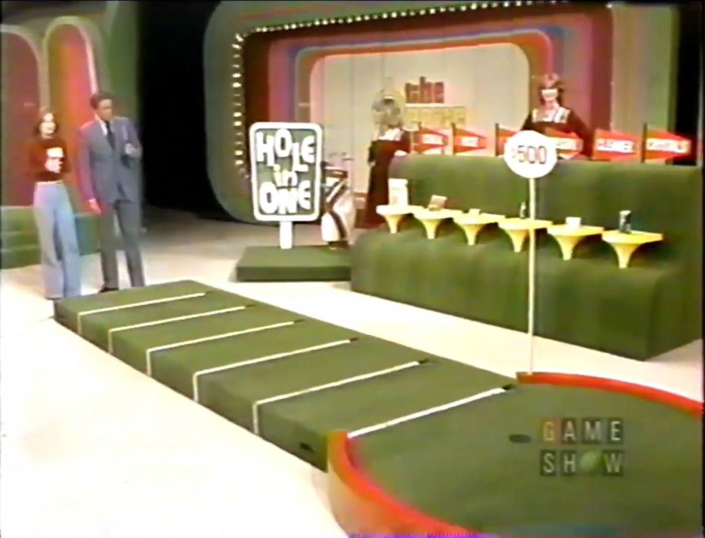 Contestants participate in the 'Hole in One' game with a putting green and flagged grocery products.