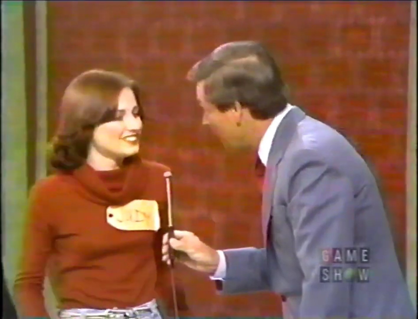 A contestant named Judy smiles while speaking to the host of a game show, wearing a red sweater and holding a microphone.