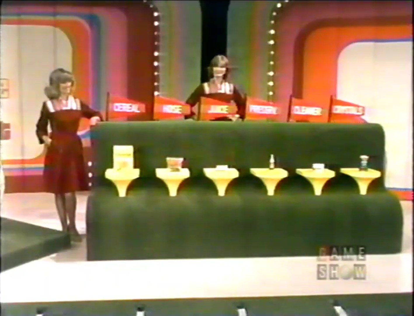 A model stands beside a stage with six grocery products displayed on yellow stands, labeled with flags indicating their prices, as part of the game 'Hole in One' on The Price is Right.