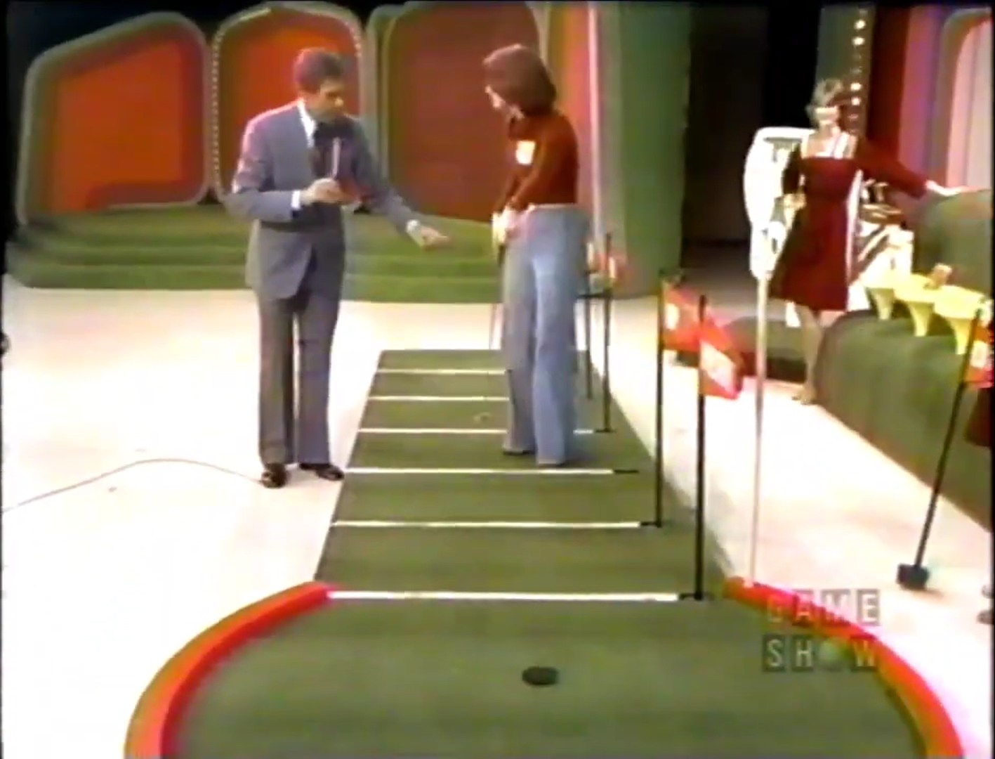 A scene from The Price is Right featuring a putting green with six lines and grocery products. A male host interacts with a female contestant, guiding her through the game while another female stands nearby.