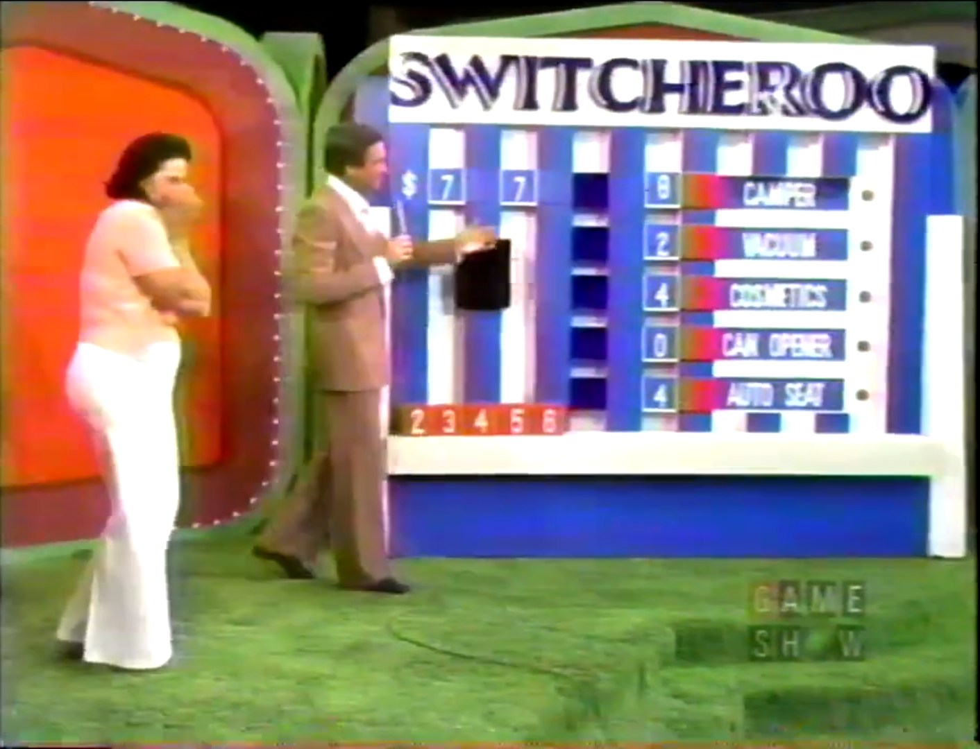 A contestant and host are at the Switcheroo game board, featuring five prize prices with missing digits, including a camper, vacuum, cosmetics set, can opener, and auto seat.