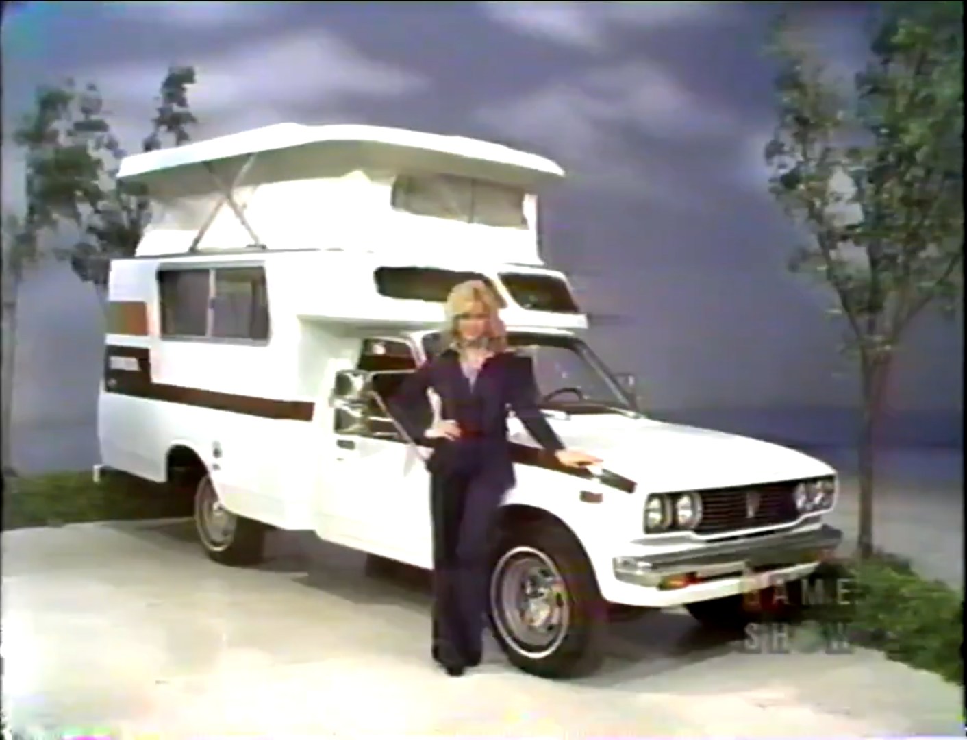 A blonde woman in a navy outfit stands next to a white camper vehicle with a raised roof, set against a backdrop of trees and a cloudy sky.