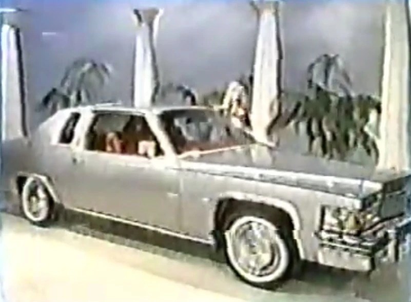 A shiny silver 1977 Cadillac Coupe de Ville showcased on The Price is Right, surrounded by decorative pillars and palm trees.
