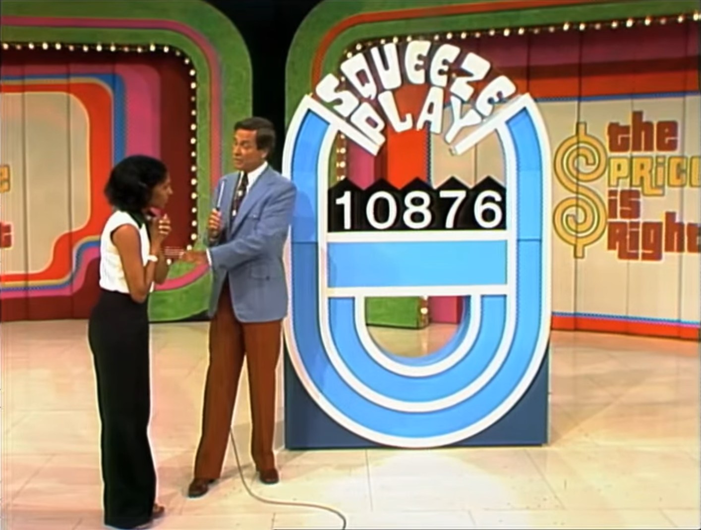 A contestant nervously discusses a game with the host in front of the Squeeze Play board displaying the incorrect price 10876.