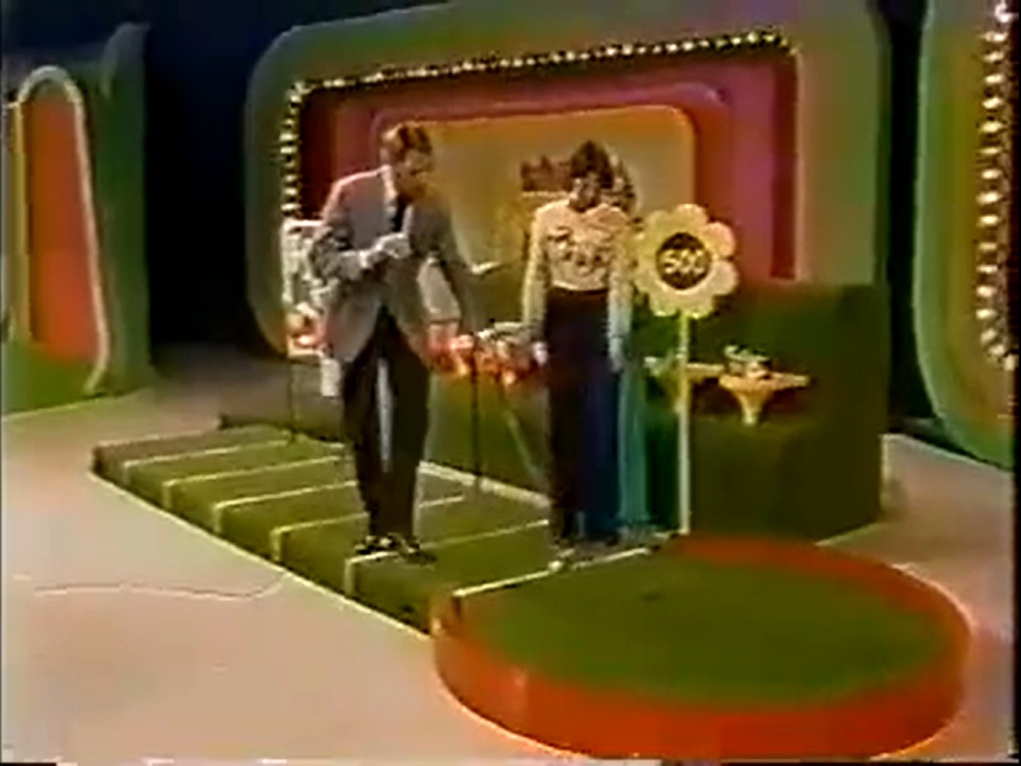 A contestant prepares to putt on the 'Hole in One' game from The Price is Right, with the host guiding them. The stage features a putting green, decorative backdrops, and a sign indicating a 0 prize.